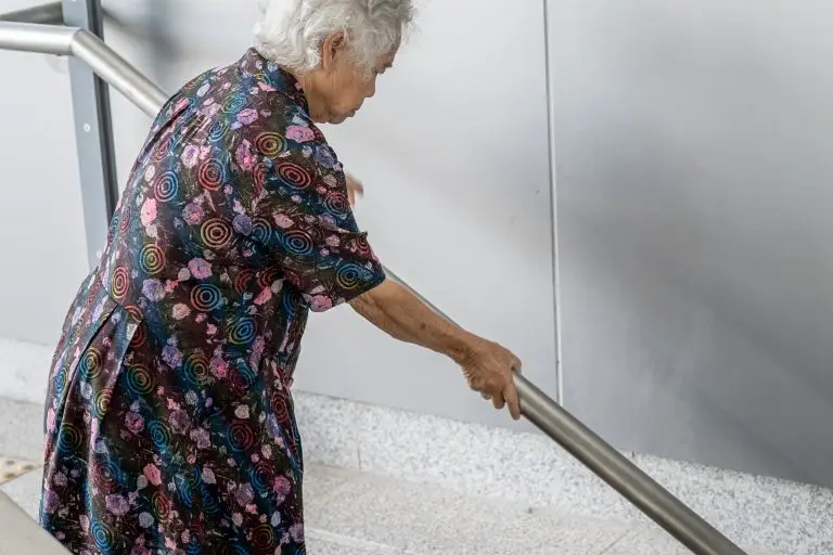 An elderly woman walks down steps with both hands on the handrail